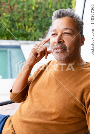 Mature man sitting on patio chair by pool, wearing brown crewneck sweater, holding hand to temple Mature man sitting on patio chair by pool, wearing brown crewneck sweater, holding hand to temple 140567366