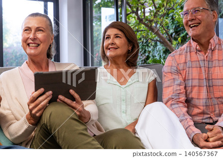 Senior woman and mature friends sitting on grey sofa holding tablet and laughing in living room Senior woman and mature friends sitting on grey sofa holding tablet and laughing in living room 140567367