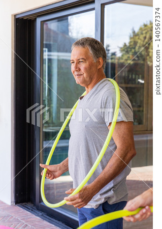 Senior man holding neon hula hoop on patio by sliding glass door in T-shirt and shorts Senior man holding neon hula hoop on patio by sliding glass door in T-shirt and shorts 140567374