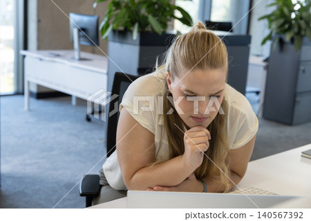 Female worker in light blouse leaning forward at office, chin on hand, focusing on laptop keyboard Female worker in light blouse leaning forward at office, chin on hand, focusing on laptop keyboard 140567392