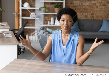 Midadult African American woman speaking and gesturing while sitting at desk in office with helmets Midadult African American woman speaking and gesturing while sitting at desk in office with helmets 140567404