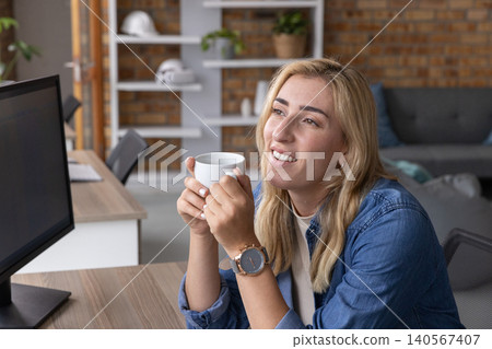 Mid-adult woman holding white ceramic mug with both hands, gazing right at monitor in office Mid-adult woman holding white ceramic mug with both hands, gazing right at monitor in office 140567407