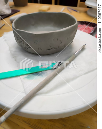 A handmade clay bowl with embossed heart and clover patterns rests on a pottery wheel, accompanied by a wooden modeling tool and a plastic scraper. A handmade clay bowl with embossed heart and clover patterns rests on a pottery wheel, accompanied by a wooden modeling tool and a plastic scraper. 140567657