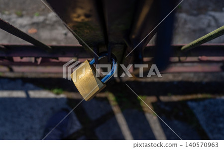 Brass padlock hanging open on weathered metal gate bar with blurred walkway below Brass padlock hanging open on weathered metal gate bar with blurred walkway below 140570963