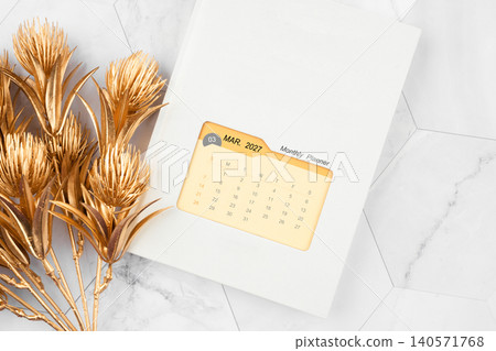 March 2027 calendar on monthly planner book with golden flower on white marble background. 140571768