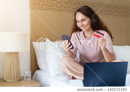 Woman shopping online with smartphone and credit card while sitting on bed Woman shopping online with smartphone and credit card while sitting on bed 140572385