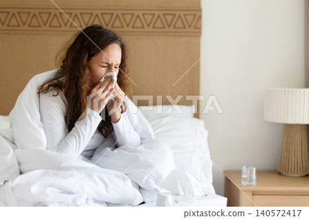 Woman sneezing into tissue while resting in bed, showing cold symptoms 140572417