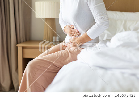 Woman holding her abdomen while sitting beside a bed, experiencing stomach pain Woman holding her abdomen while sitting beside a bed, experiencing stomach pain 140572425
