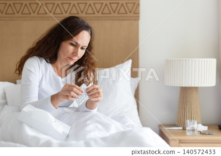 Sick woman sitting in bed examining blister pack of pills Sick woman sitting in bed examining blister pack of pills 140572433