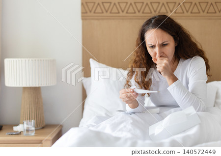 Sick woman coughing while checking thermometer in bed, illustrating respiratory illness, Sick woman coughing while checking thermometer in bed, illustrating respiratory illness, 140572449