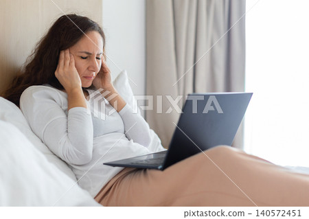 Woman resting in bed with laptop while suffering from headache Woman resting in bed with laptop while suffering from headache 140572451