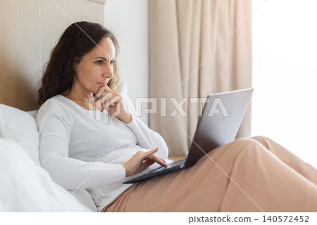 Thoughtful adult woman working on laptop while sitting in bed Thoughtful adult woman working on laptop while sitting in bed 140572452