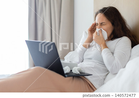 Sick woman sneezing into tissue while using laptop in bed, representing illness Sick woman sneezing into tissue while using laptop in bed, representing illness 140572477