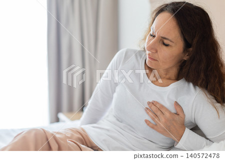 Woman sits on bed holding hand to chest with pained expression Woman sits on bed holding hand to chest with pained expression 140572478