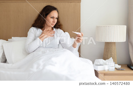 Concerned woman checking thermometer while sitting in bed, monitoring fever symptoms Concerned woman checking thermometer while sitting in bed, monitoring fever symptoms 140572480