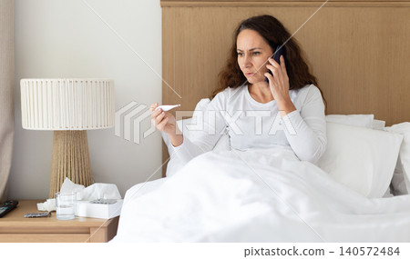 Woman talking on smartphone while checking thermometer in bed Woman talking on smartphone while checking thermometer in bed 140572484