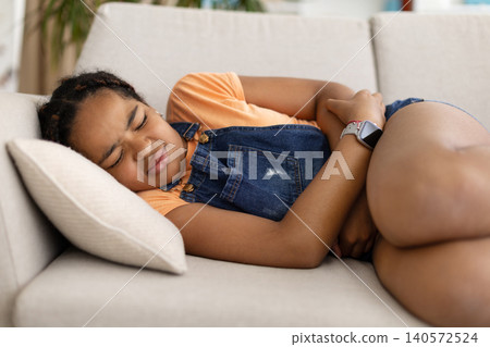 Black teenage girl lying on sofa holding her stomach and showing discomfort 140572524