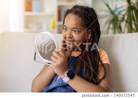 Black teenage girl looking at her reflection in handheld mirror at home Black teenage girl looking at her reflection in handheld mirror at home 140572545