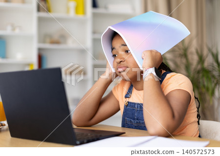 Black teenage girl covering head with notebook while studying online with laptop Black teenage girl covering head with notebook while studying online with laptop 140572573