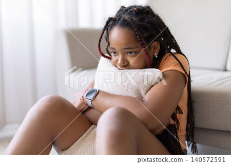 Black teenage girl sitting on floor hugging cushion at home, suffering depression Black teenage girl sitting on floor hugging cushion at home, suffering depression 140572591