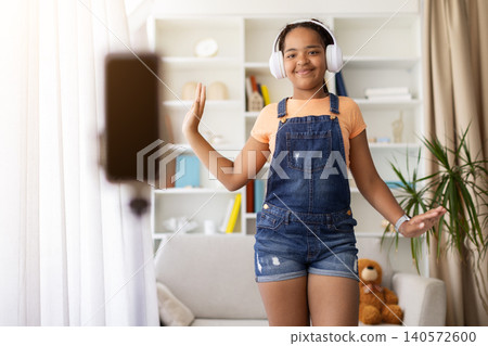 Black teenage girl dancing and recording video at home while wearing headphones 140572600