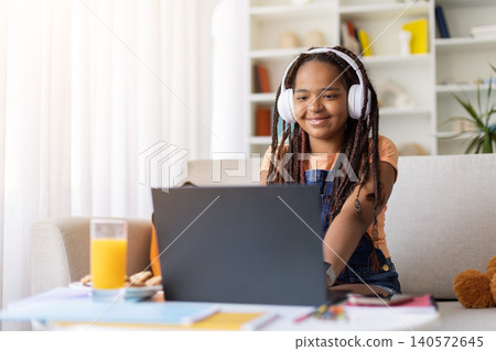Black teenage girl wearing headphones using laptop while studying at home Black teenage girl wearing headphones using laptop while studying at home 140572645