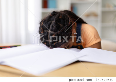 Black teenage girl rests head on open notebook at desk, appearing exhausted Black teenage girl rests head on open notebook at desk, appearing exhausted 140572650