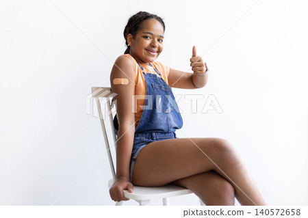 Black teenage girl sitting on chair showing bandage on her arm and giving thumbs up Black teenage girl sitting on chair showing bandage on her arm and giving thumbs up 140572658