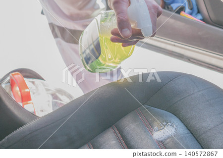 Close up of a professional male detailer spraying a chemical cleaning solution from a bottle onto a dirty fabric car seat during an automotive interior detailing Close up of a professional male detailer spraying a chemical cleaning solution from a bottle onto a dirty fabric car seat during an automotive interior detailing 140572867