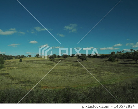 A landscape of grasslands and rolling hills unfolds outside the train window—a tranquil scene of land beneath a blue sky. 140572954