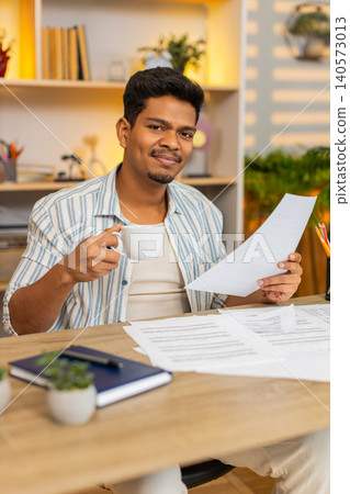 Indian man on home table drinking coffee and checking documents during focused work in living room Indian man on home table drinking coffee and checking documents during focused work in living room 140573013