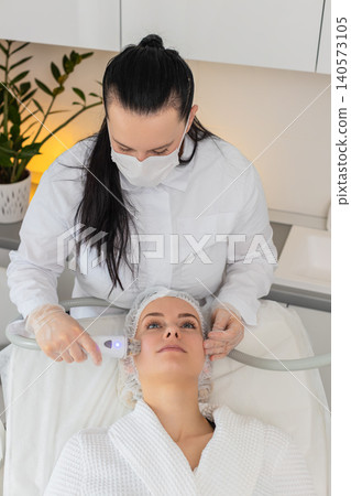 Beautician makes woman RF lifting cosmetology procedure of face and cheek area, collagen mask at spa Beautician makes woman RF lifting cosmetology procedure of face and cheek area, collagen mask at spa 140573105
