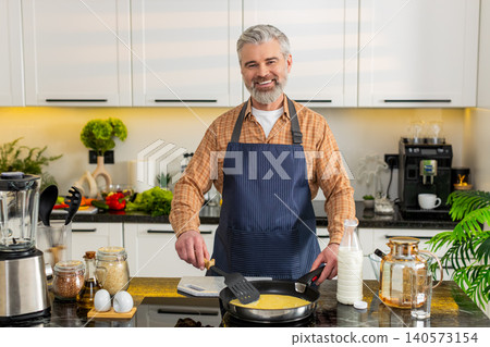 Middle-aged man pouring raw omelet mix into frying pan in home kitchen for healthy diet breakfast. Middle-aged man pouring raw omelet mix into frying pan in home kitchen for healthy diet breakfast. 140573154