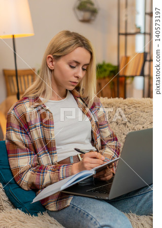 Adult woman writing financial notes in notebook while looking at laptop screen at home with focus Adult woman writing financial notes in notebook while looking at laptop screen at home with focus 140573197