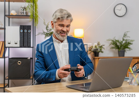 Middle-aged businessman gamer holds joystick wins laptop race shouting victory with celebration pose Middle-aged businessman gamer holds joystick wins laptop race shouting victory with celebration pose 140573285