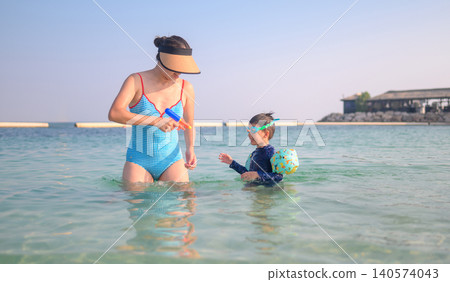 Asian woman in swimsuit playing with her 4-year-old son in clear beach water Asian woman in swimsuit playing with her 4-year-old son in clear beach water 140574043