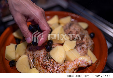 Hand placing black olives into a traditional moroccan earthenware tagine pot, preparing chicken and chunked potatoes for cooking a savory, fresh meal Hand placing black olives into a traditional moroccan earthenware tagine pot, preparing chicken and chunked potatoes for cooking a savory, fresh meal 140574338