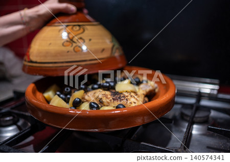 Person cooking traditional moroccan chicken tagine, simmering with potatoes and olives inside a clay pot on a gas stove, preparing a flavorful and healthy meal 140574341