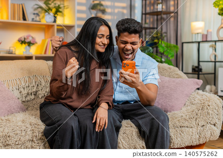 Indian couple man and woman sitting on home sofa celebrating victory on smartphone after good news Indian couple man and woman sitting on home sofa celebrating victory on smartphone after good news 140575262