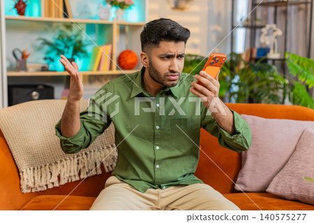 Indian man on home sofa arguing on smartphone speaker call with stop gesture and nervous reaction Indian man on home sofa arguing on smartphone speaker call with stop gesture and nervous reaction 140575277