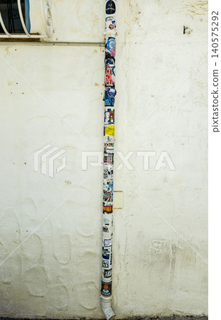 ALTEA, SPAIN - JUNE 17, 2026. A vertical drainpipe runs along a white stucco wall. The pipe is covered with colorful stickers and street art. This scene shows urban art in an everyday city environment ALTEA, SPAIN - JUNE 17, 2026. A vertical drainpipe runs along a white stucco wall. The pipe is covered with colorful stickers and street art. This scene shows urban art in an everyday city environment 140575292