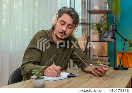 Middle-aged man listening audiobook headphones taking notes in notebook deep focused study at table Middle-aged man listening audiobook headphones taking notes in notebook deep focused study at table 140575358