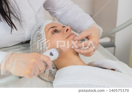 Woman receives RF lifting facial massage therapy by cosmetologist using professional equipment 140575370
