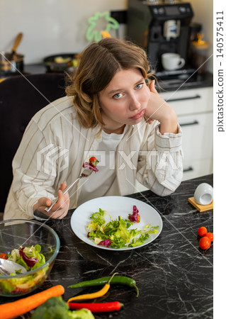 Young woman feeling upset while eating healthy salad during strict diet discipline in home kitchen 140575411