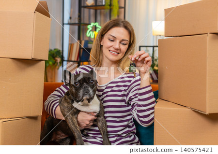 Middle-aged woman with dog raises keys shows victory pose sits by boxes after move to new place calm Middle-aged woman with dog raises keys shows victory pose sits by boxes after move to new place calm 140575421