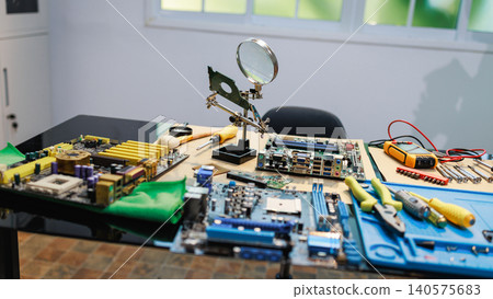 Computer repair workspace with circuit boards, motherboard parts, hand tools and a magnifying glass on a table in a bright room. Computer repair workspace with circuit boards, motherboard parts, hand tools and a magnifying glass on a table in a bright room. 140575683