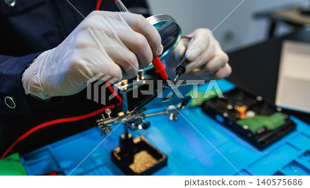 Technician in gloves testing a hard drive circuit board with multimeter probes on a workbench, showing electronics repair, data recovery, and careful hands-on maintenance. 140575686
