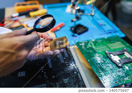 Close-up of hands inspecting a computer processor with a magnifying glass on a workbench with circuit boards and repair tools. Close-up of hands inspecting a computer processor with a magnifying glass on a workbench with circuit boards and repair tools. 140575697