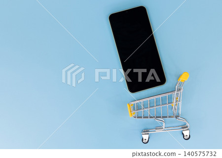 Minimalist scene with smartphone and shopping cart. Flat lay on solid blue background. Modern device beside miniature retail cart. Suggests online shopping or e-commerce concept Minimalist scene with smartphone and shopping cart. Flat lay on solid blue background. Modern device beside miniature retail cart. Suggests online shopping or e-commerce concept 140575732