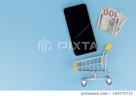 Minimalist scene with smartphone and shopping cart. Flat lay on solid blue background. Modern device beside miniature retail cart. Suggests online shopping or e-commerce concept Minimalist scene with smartphone and shopping cart. Flat lay on solid blue background. Modern device beside miniature retail cart. Suggests online shopping or e-commerce concept 140575733